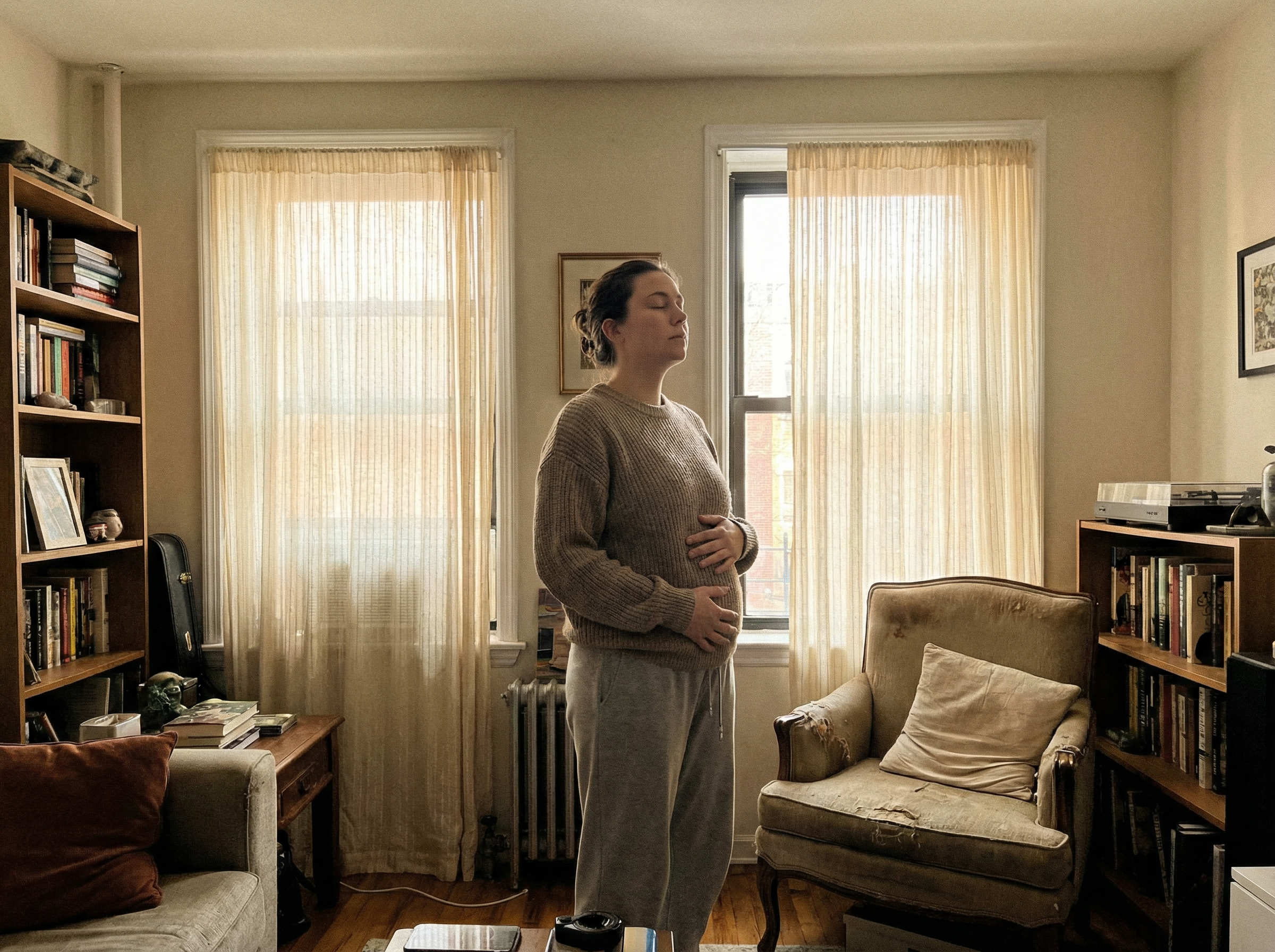 Person doing standing breathing exercise in apartment living room, hands over lower abdomen, morning light