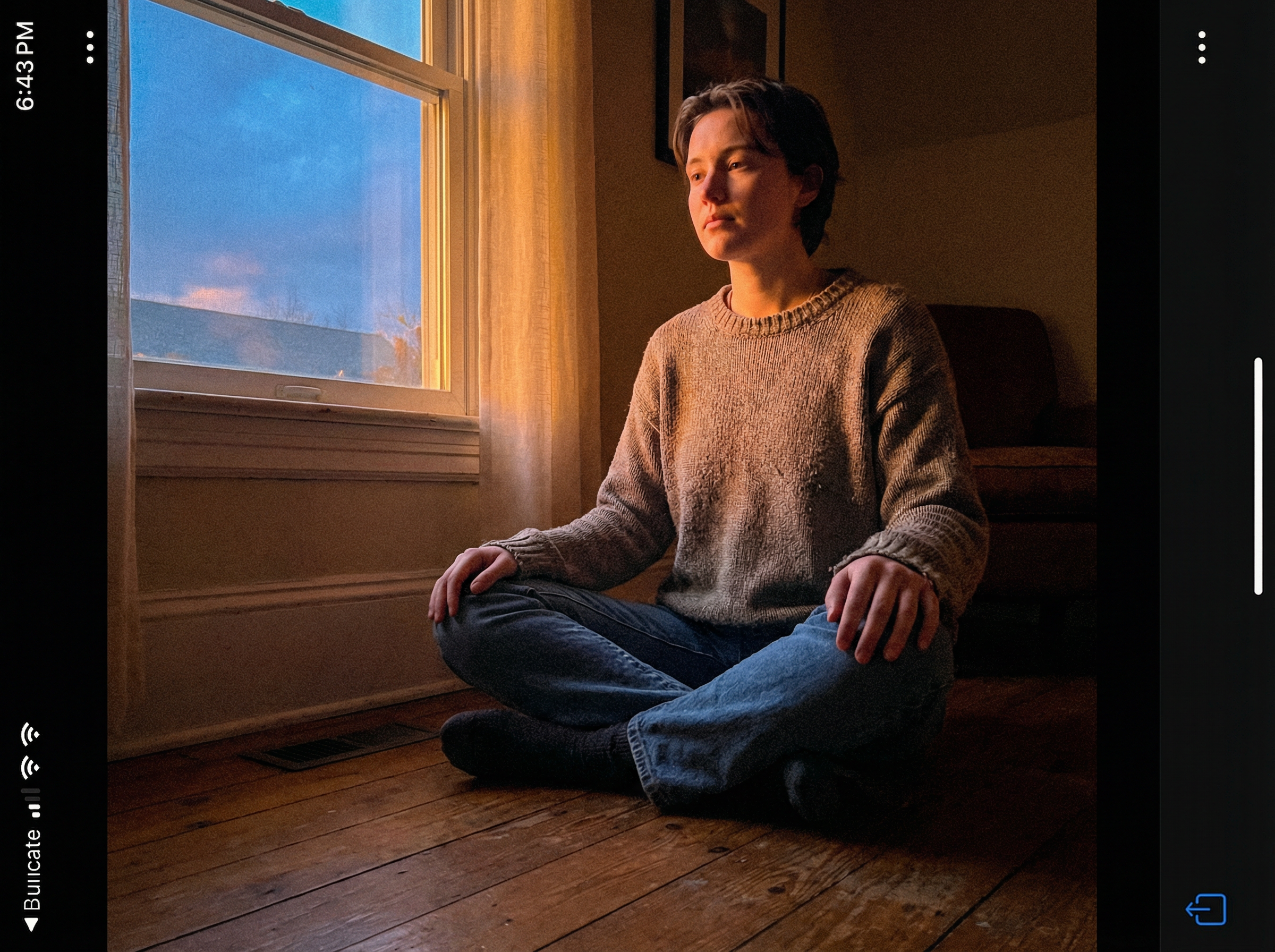Person sitting cross-legged on wooden floor near window at dusk, calm and meditative, golden light