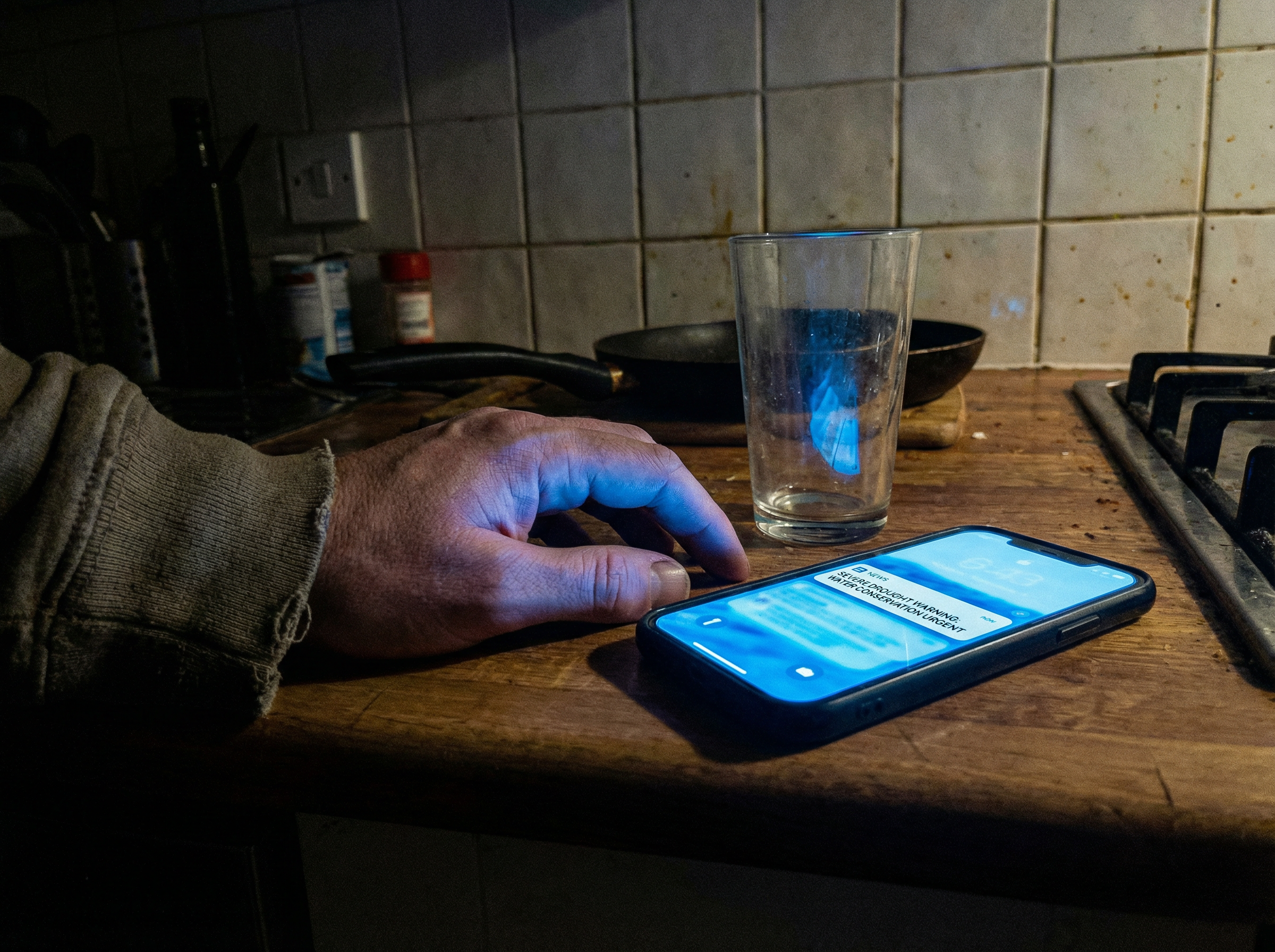 A dimly lit kitchen counter at 3am, a half-empty glass of water next to a glowing smartphone showing a news alert about drought, casting a harsh blue light on a tired hand resting on the counter.